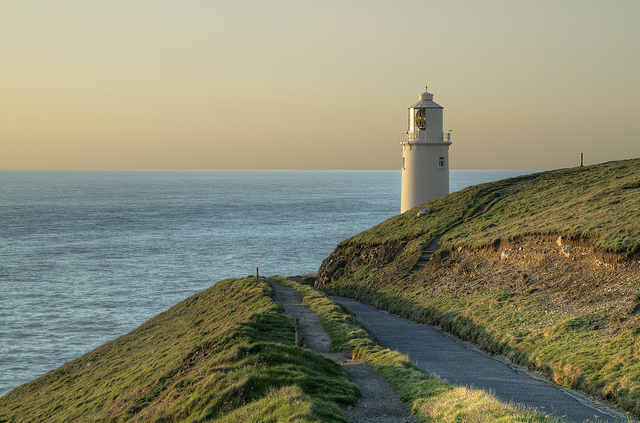 White lighthouse located on a coastal clifeside coastal path with green hills, blue water and a creamy sky at sunset