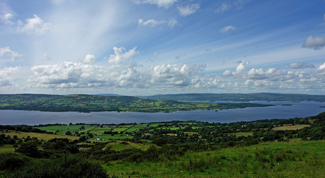 Landscape image of a blue lake amongst green hills and a cloudy blue sky