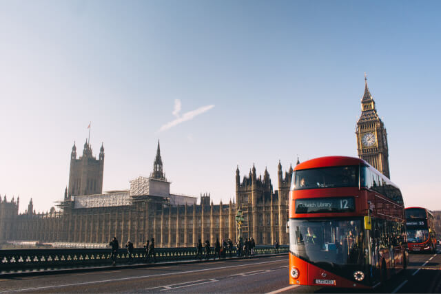 A view of the Palace of Westminster from the Westminster Bridge, with Big Ben visible in the background and an iconic double-decker bus in the foreground.
