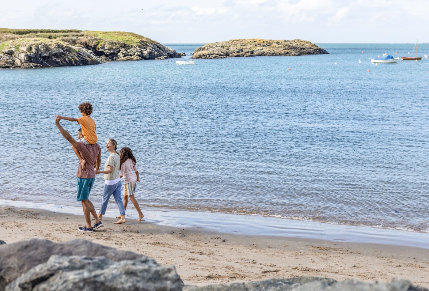 Family walking along beach with sea in background. Son on dad's shoulders while mum holds daughter's hand.