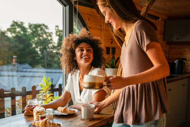 Female couple enjoying breakfast in log cabin. One girl is standing with coffee mug in hand