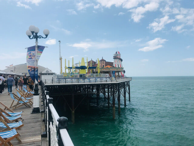 Brighton Palace Pier boardwalk with rides and amusements in the near distance and the sea on the right, blue skies overhead.