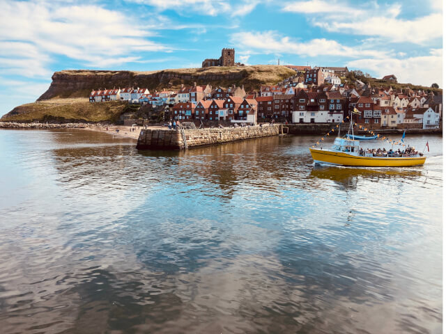 Boat sails across Whitby Harbour with the ruined Whitby Abbey in background