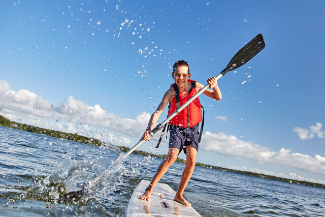 A young boy stood on a board in the water holding a paddle