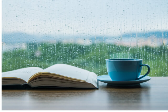 Open book and a blue cup and saucer placed on a table in front of a rainy window