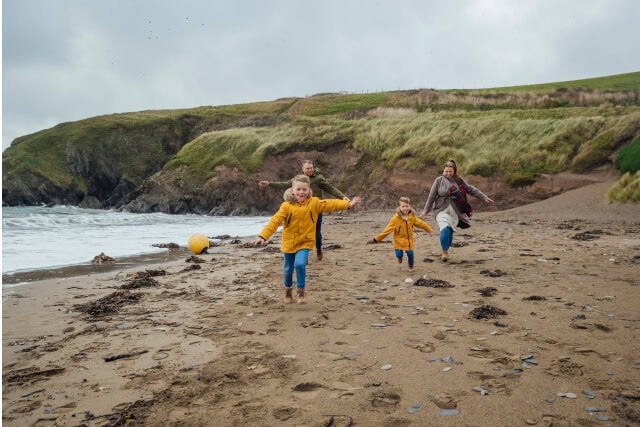 Family with children running on the beach alongside the sea, with rugged, grassy cliffs in the background