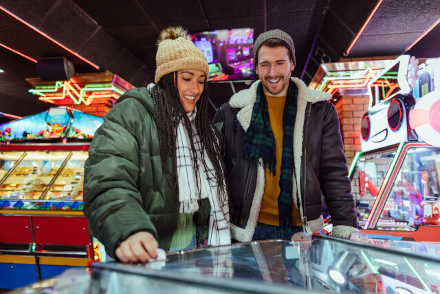 Male female couple both smiling, playing an arcade game together, wearing winter clothes