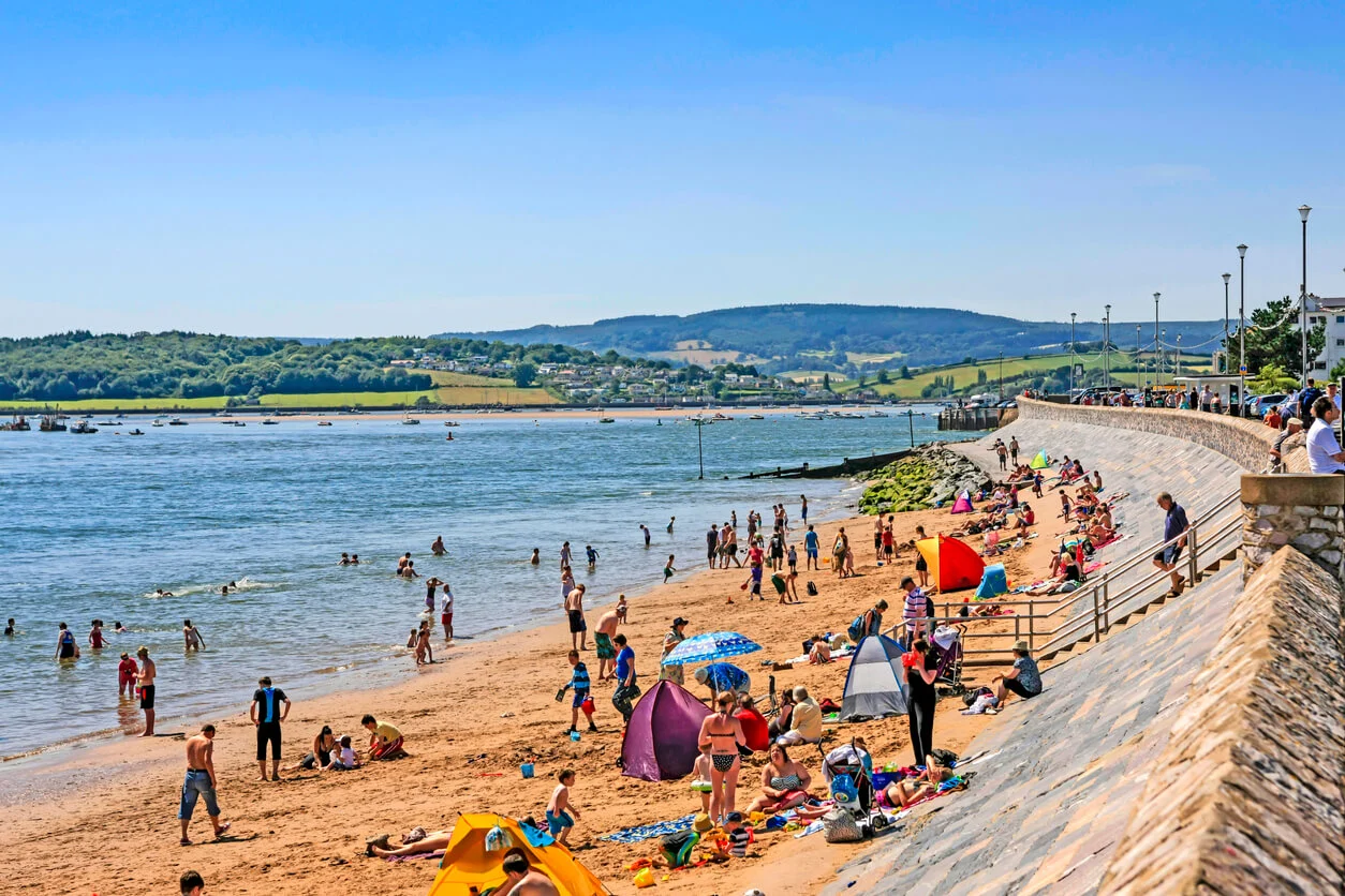 a vibrant beach scene in Exmouth, England. The beach is bustling with activity, with people enjoying the sun, sand, and sea. Families and groups are spread out along the sandy shore, some under colorful umbrellas and tents, while others are playing in the water. The beach is bordered by a curved sea wall with steps leading down to the sand, and there are people walking along the promenade above. The water is calm, and in the distance, you can see a lush, green landscape with rolling hills and scattered houses, creating a picturesque backdrop for this lively coastal scene.