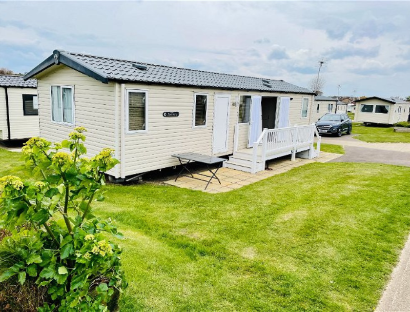 A light beige mobile home with a black roof and white trim at Primrose Valley Caravan Park, available through UK Caravans 4 Hire. The home features a small white deck with railings and steps leading to a paved patio area with a folding table. Green plants with yellowish flowers are visible in the foreground. The setting includes well-maintained green lawns, other mobile homes, and a car parked nearby under a cloudy sky, showcasing a pleasant and organised holiday environment.