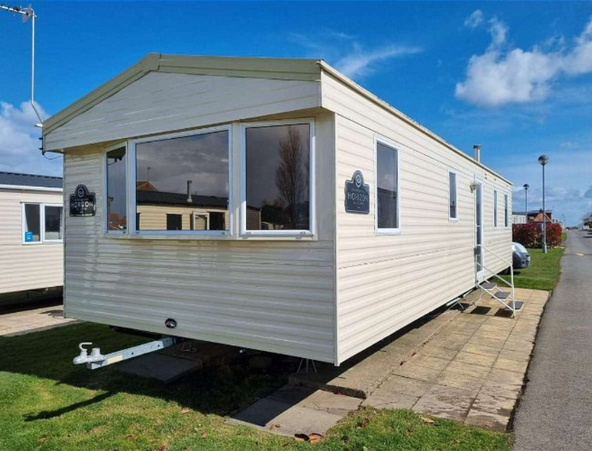 A light beige mobile home with large windows and white trim, located at Primrose Valley Caravan Park. The home has a small set of metal steps leading to the entrance and is situated on a paved and grassy area. The bright, clear blue sky adds to the inviting atmosphere. Other mobile homes and a road are visible in the background, indicating a well-organized and maintained park setting. This accommodation option, available through UK Caravans 4 Hire, offers a comfortable and convenient stay in a picturesque environment.