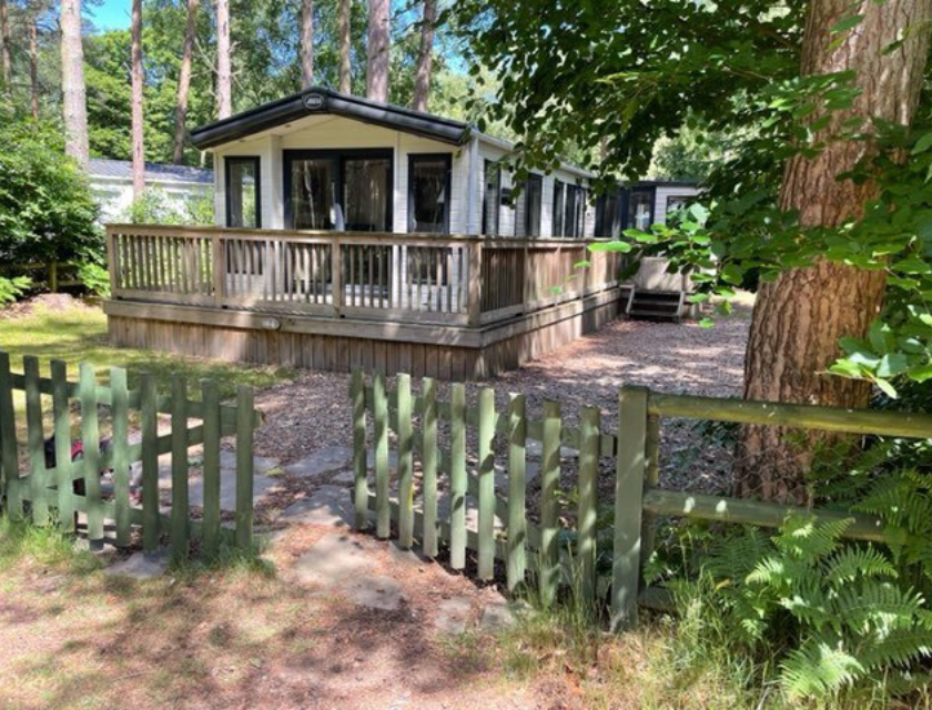 A cosy beige mobile home with a dark roof and large windows, located at Primrose Valley Caravan Park and available through UK Caravans 4 Hire. The home features a spacious wooden deck with railings, set in a wooded area surrounded by tall trees and lush greenery. A green picket fence and a stone pathway lead to the entrance, creating a welcoming and serene atmosphere. The natural setting highlights the tranquil and secluded environment, perfect for a relaxing getaway.