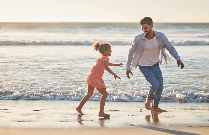 Accompanying image for Parkdean Resorts in England for families to consider. | Image is of a child and father on beach running in ocean together together for body movement wellness, exercise and healthy development. Dad having fun and playing with girl kid at sea ocean outdoor summer holiday