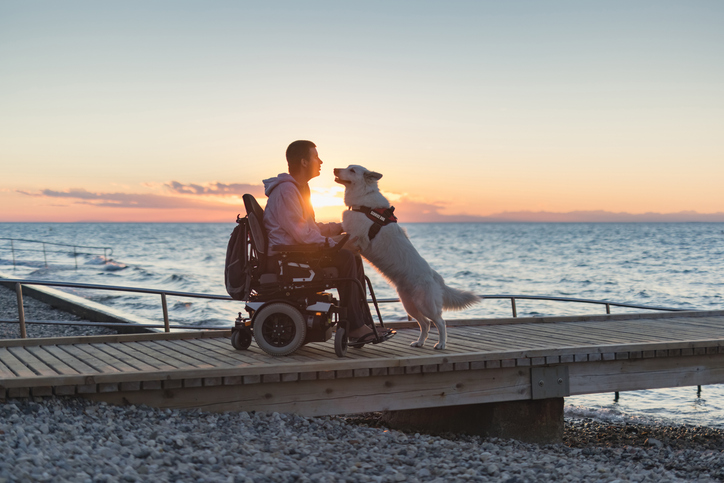 Sandy Bay Beach is accessible for people with low mobility | Image of a a man with disability with his service dog at sunset using electric wheelchair.