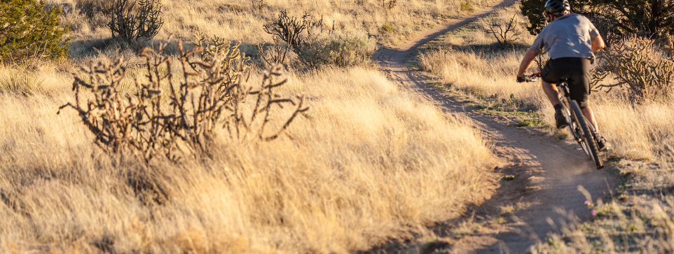 Single rider turning a corner on a bike on a cycling trail. The trail is dusty and the picture is taken with a sunrise/set in the background.