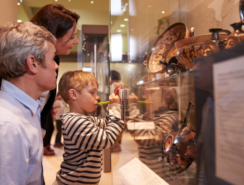 Family observing and interacting with historical artifacts in a museum display case, with a young boy focused on the exhibit. Example of a rainy day thing to do in Cornwall for families.