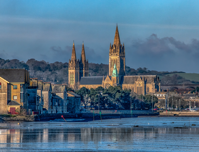 Shot taken across water of Truro skyline with Truro Cathedral visible and surrounding stone buildings. The sky is grey and cloudy.
