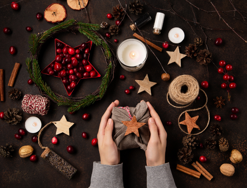 Hands wrapping a present in festive, natural materials like twine and star-shaped ornaments. Surrounded by candles, pine cones, and a wreath, it creates a cozy Christmas atmosphere.