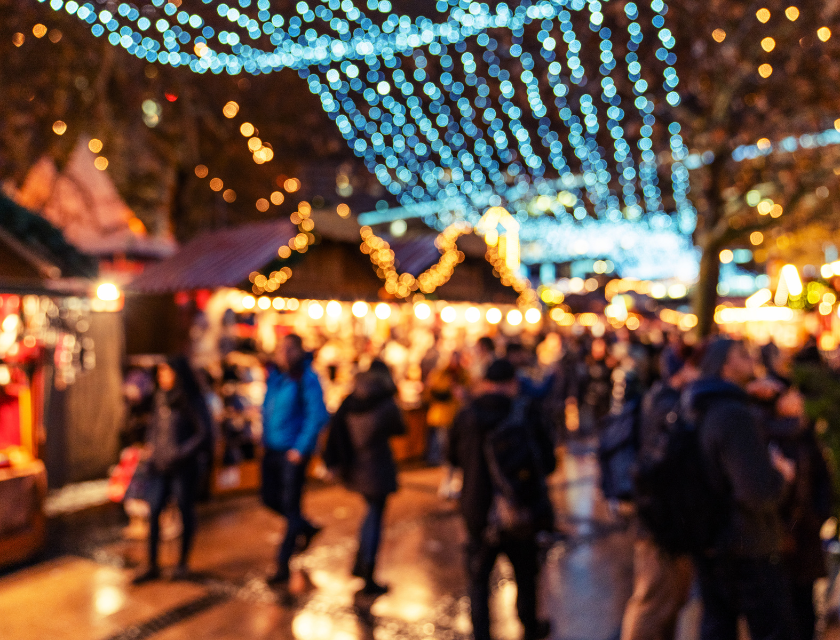 A blurry photo of a busy Christmas market at night, with glowing festive lights hanging above.